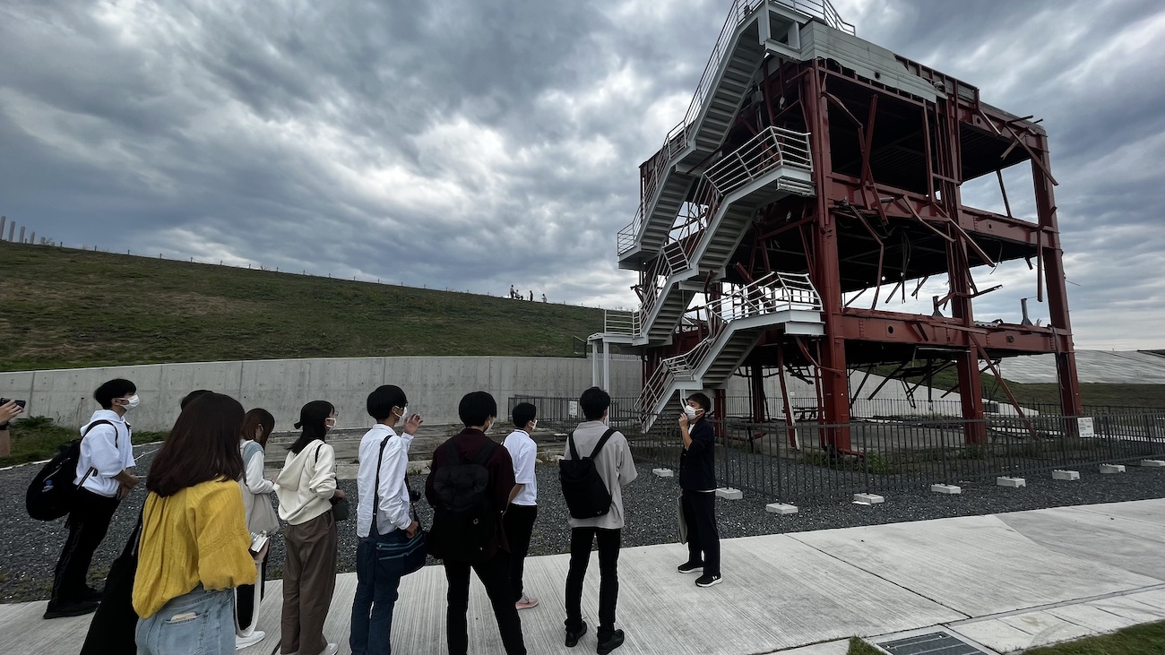被災した赤い鉄骨の建物の前で、マスクをした大学生たちがガイドの説明を受けて真剣に耳を傾けている、震災遺構を見学している様子を示した写真