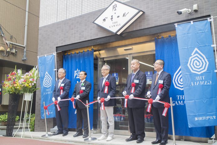 グレーのタイル張りの建物の入り口に「座・ガモール」と書かれたひし形の看板が設置されており、青いのぼりや垂れ幕が設置された店舗前でスーツ姿の男性5人が白手袋をつけてテープカットを行っている写真