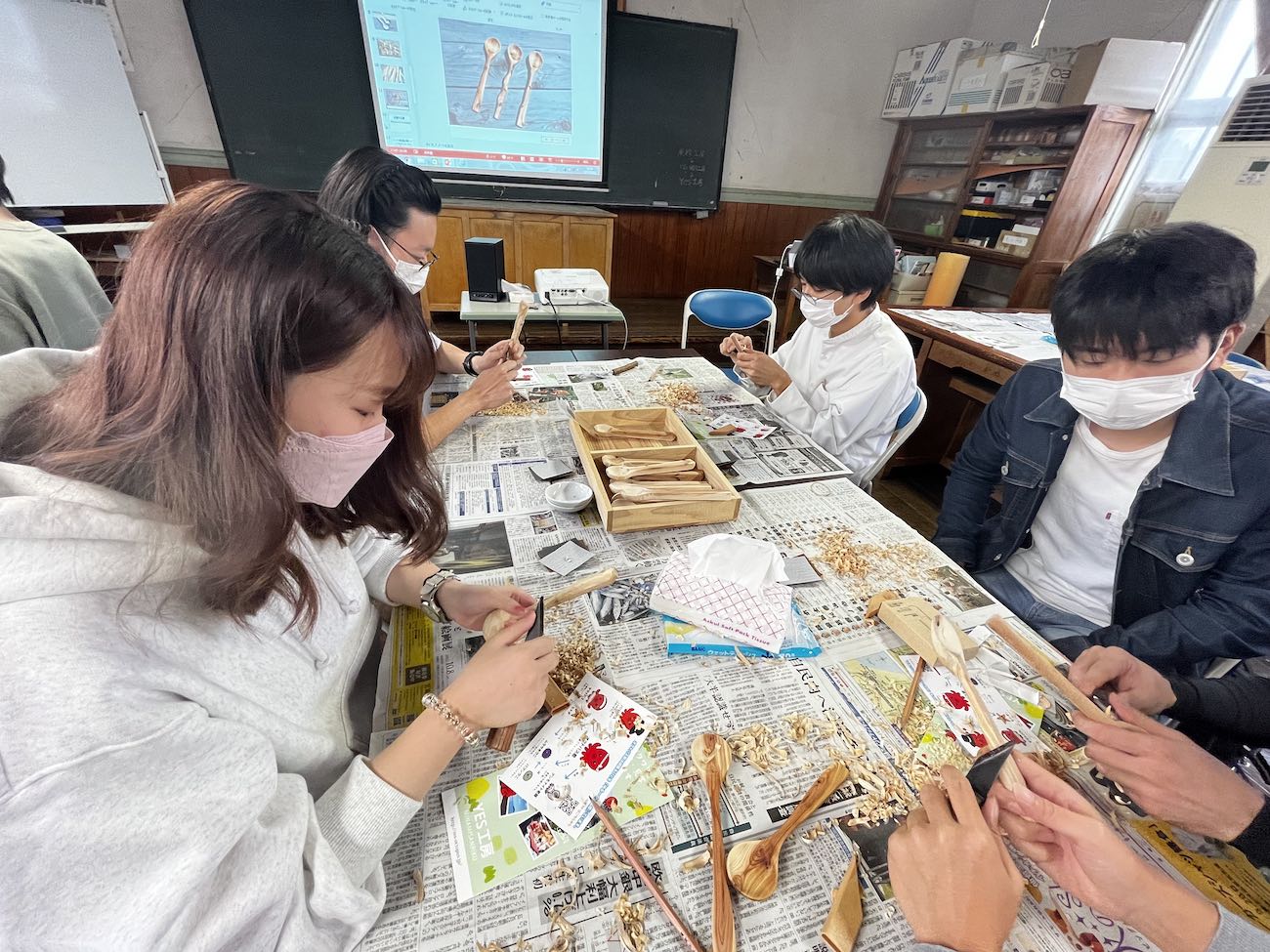 テーブルに新聞紙を敷き、大学生たちが木のスプーンを彫っている手元に集中している木工体験の様子の写真