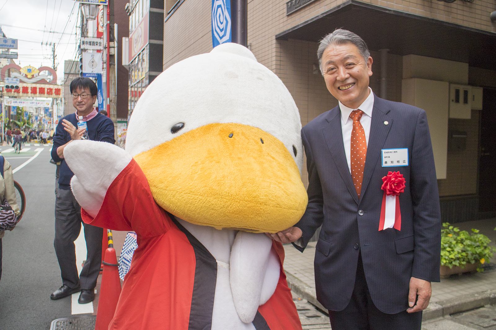 巣鴨のゆるキャラ・すがもんと最知副町長が屋外で商店街を背景に笑顔で写っている写真