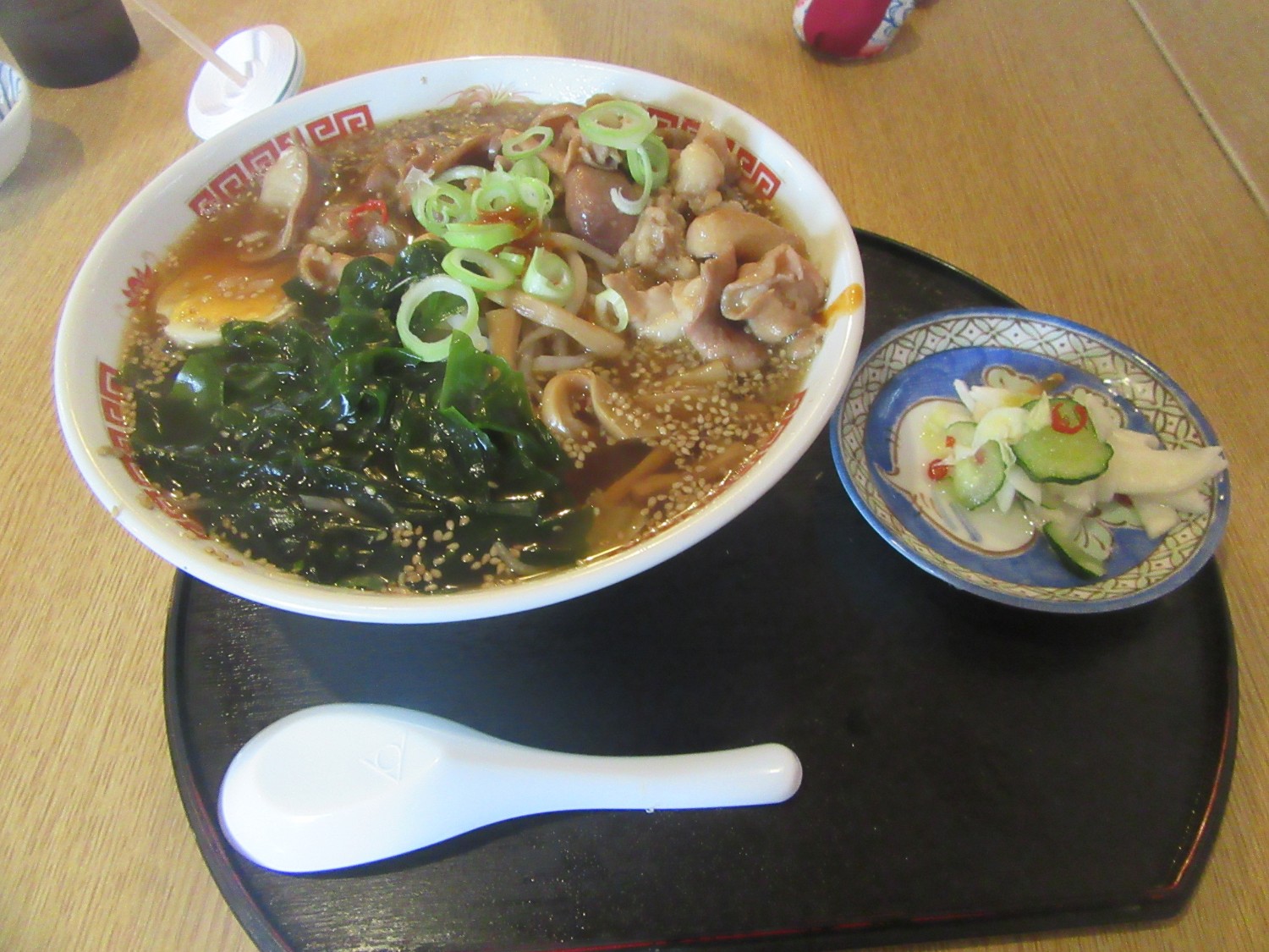 大量のわかめとネギ、煮卵、肉などがのった醤油ベースのラーメンと、きゅうりと白菜の浅漬けがお盆に並べられたラーメンの写真
