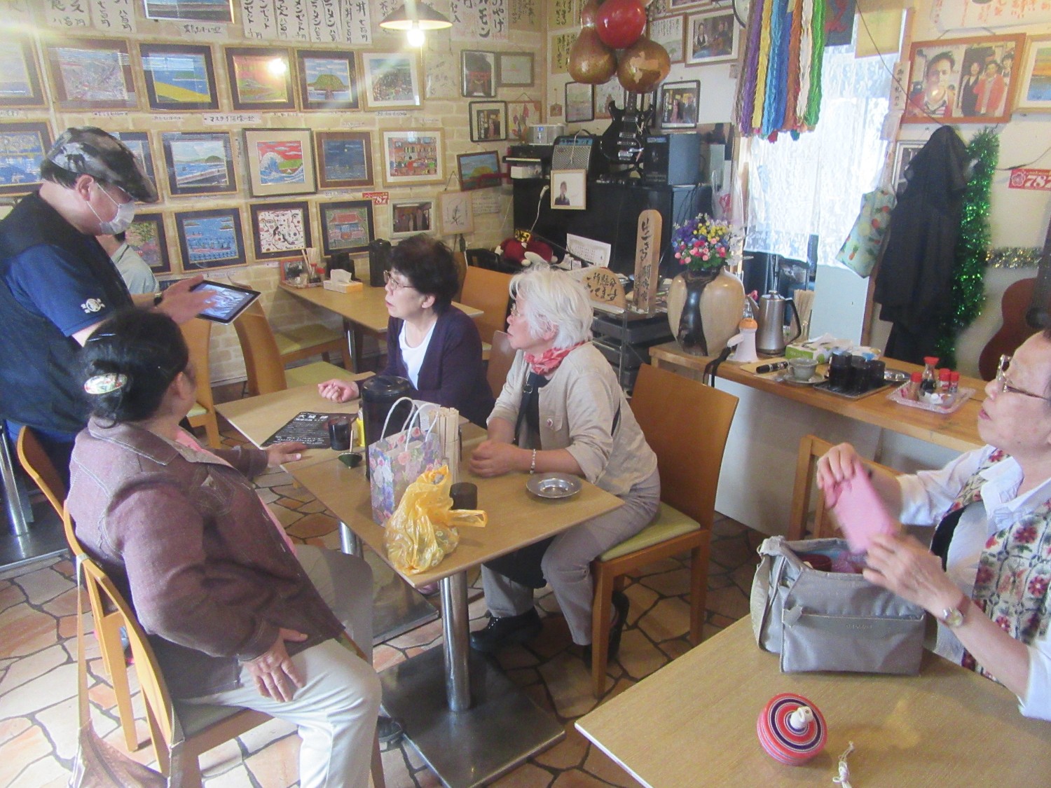 店内のテーブル席に高齢の女性たち数人が座り、ハンチング帽子をかぶった店主の渡辺さんがマスク姿でタブレットを操作しながら接客している様子の写真
