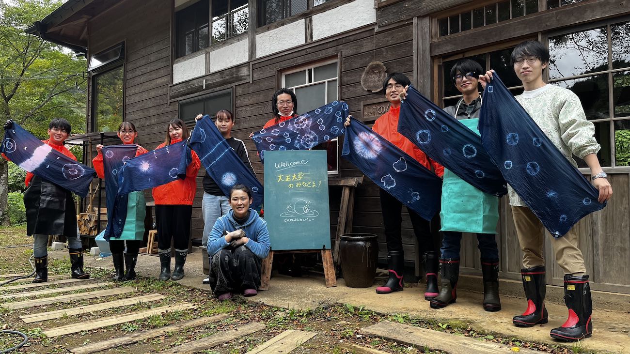 木造の古民家の前で、藍染め体験を終えた大学生たちが長靴とエプロン姿で青い染物を広げて見せながら横一列に並んでいる集合写真