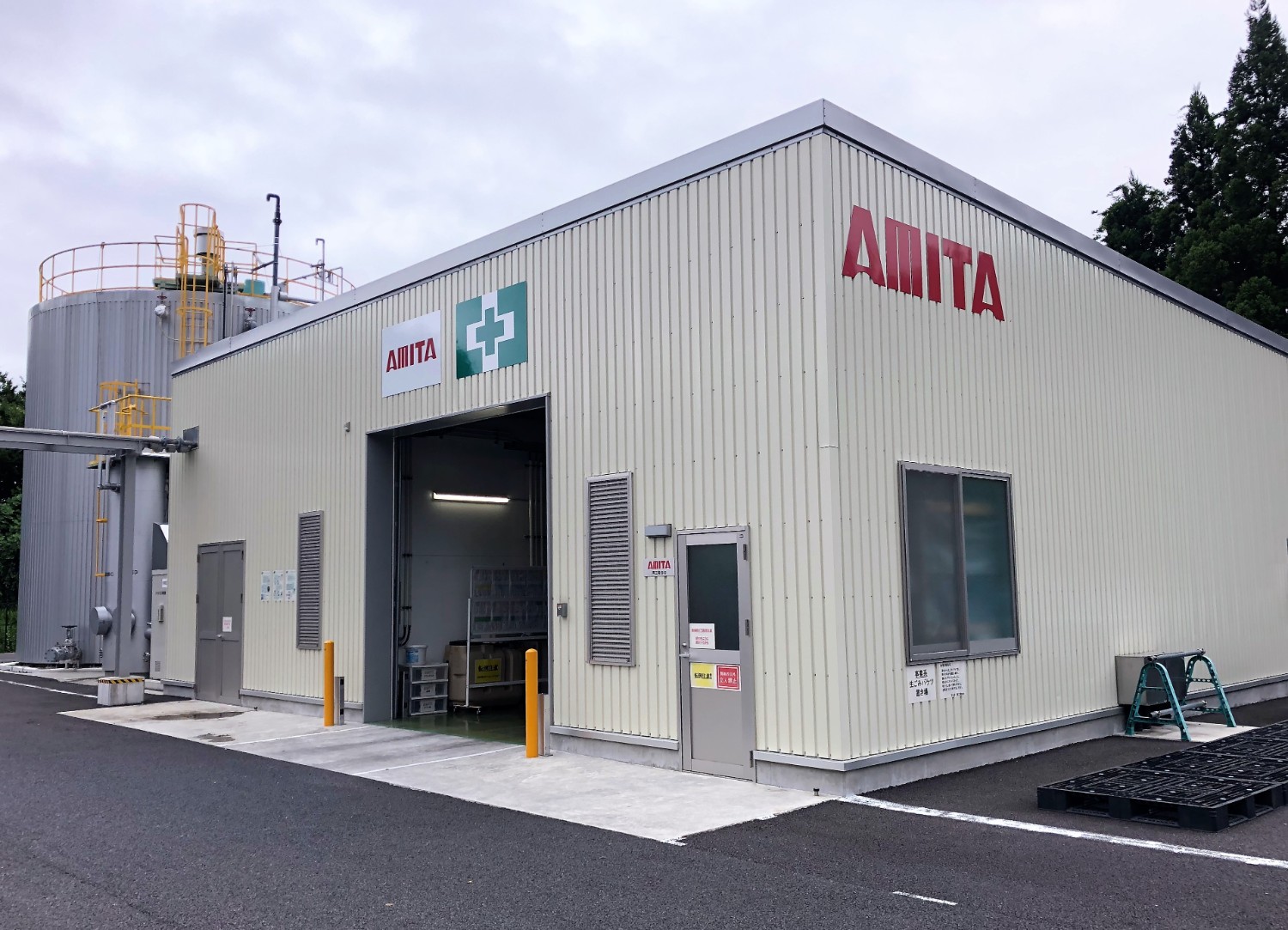 奥に銀色の大型タンクがあり、手前に赤い「AMITA」のロゴと緑の十字マークが掲示されているベージュ色の建物が建っている写真