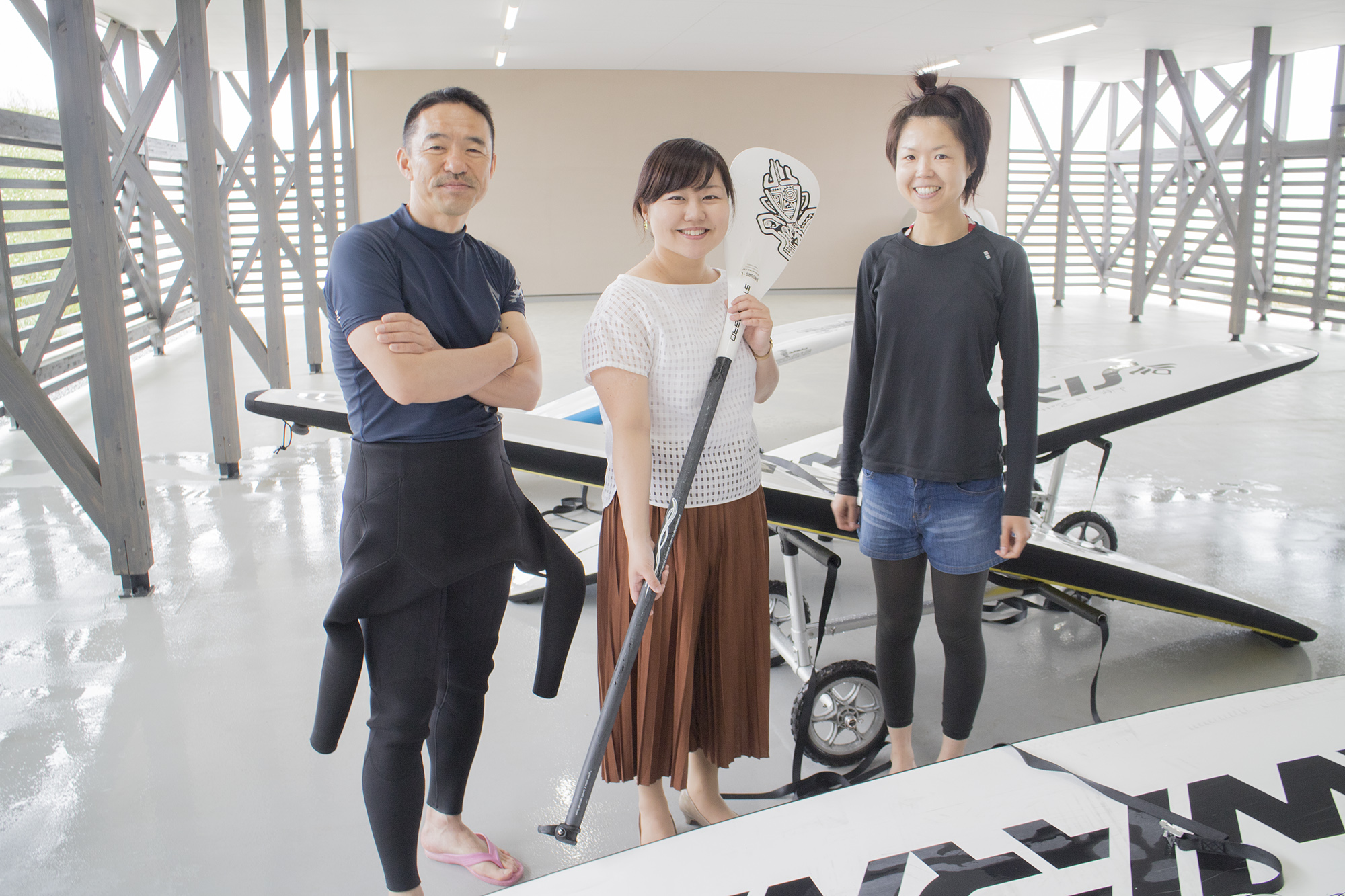 白を基調とした広々とした室内にスタンドアップパドルボード用のボードが並べられ、その間に、ウエットスーツを腰まで着用し腕組みをした男性、パドルを持ったリポーターの女性と黒い長袖とショートパンツ姿の女性が3人並んで立っている写真