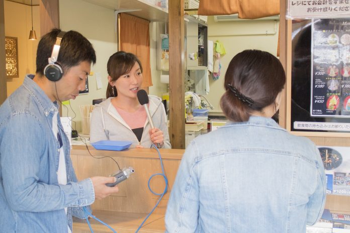 飲食店のカウンター越しにマイクを持って話をしている女性と、ヘッドホンをして機材を持った男性、後ろ姿の女性が写っている写真