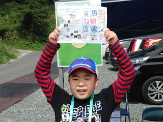 赤と黒のボーダーの服を着た少年が笑顔でスタンプが押された紙を頭の上に掲げて見せており、青い帽子をかぶっている写真