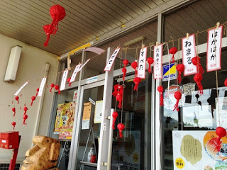 建物の入口に赤い布製の飾りと妖怪の名前が書かれた札がたくさん吊るされており、郵便ポストや案内ポスターも見える写真