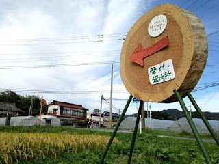 大きな木の年輪を模した看板に赤い矢印と「受付・案内所」と書かれており、周囲に田んぼと電線がある田舎道の風景の写真