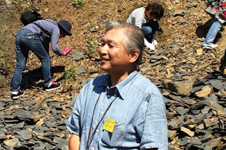 化石発掘現場で多くの参加者が作業している中、胸に名札を付けた講師の男性が笑顔で立っている様子の写真