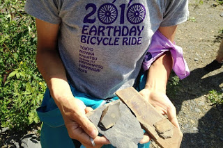 「EARTHDAY BICYCLE RIDE」と書かれたTシャツを着た人が手に石片と金属製の道具を持ち、見つけた化石を見せている写真
