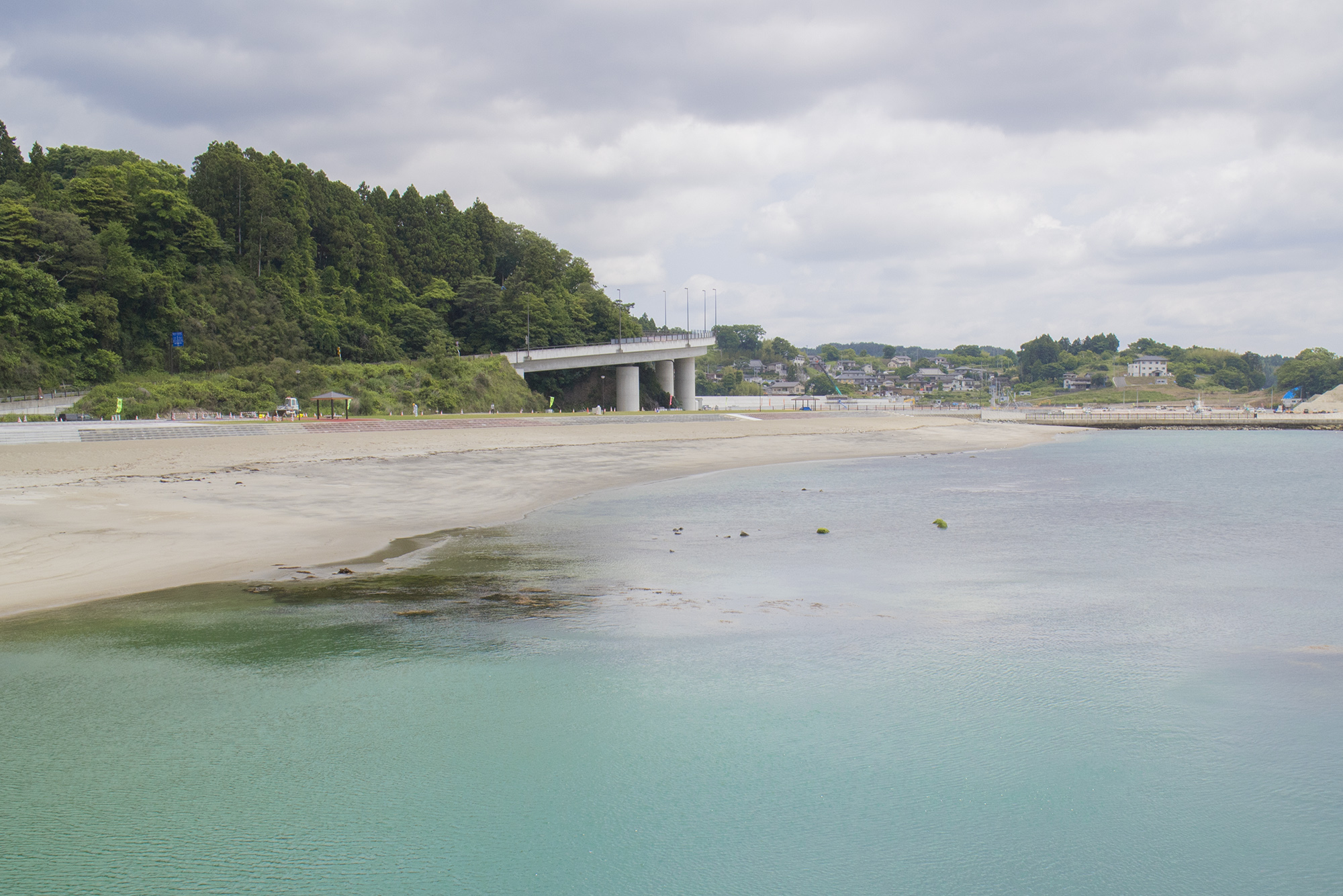 緑豊かな森の横にかかる高架道路の下に広がる、白い砂浜と透き通ったエメラルドグリーンの海、その奥には小さな町並みが広がる穏やかな海辺の風景を写した写真