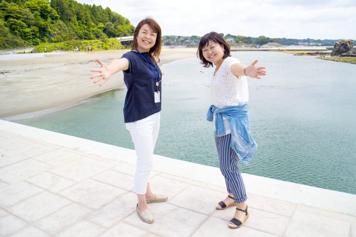 左手に砂浜が広がり、奥に森や住宅が見える海辺を背景に2人の女性が笑顔で両手を広げて立っている写真