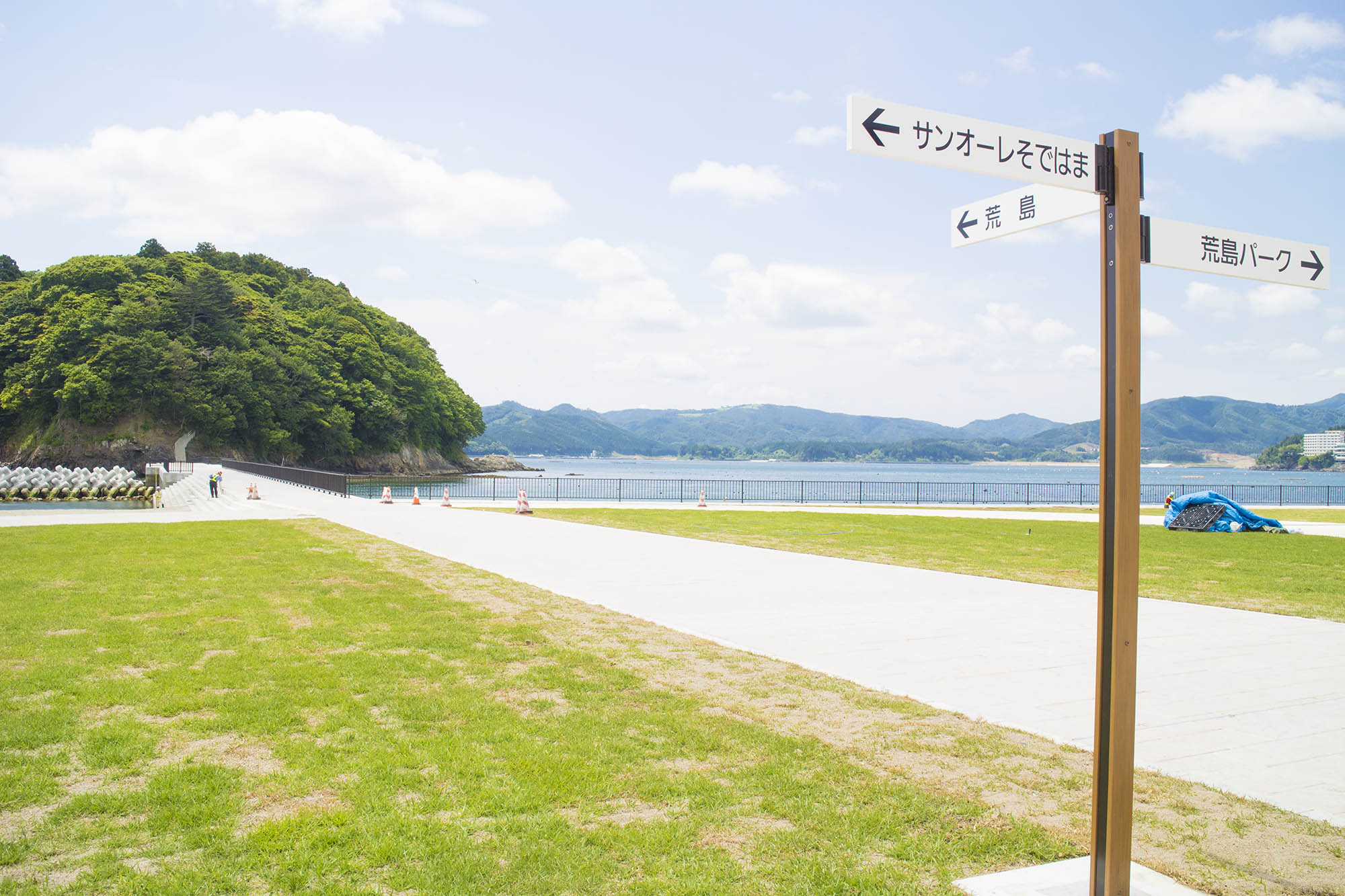 海辺の公園に設置された案内標識が中央に写っており、背景には穏やかな海と緑豊かな山々が広がっている風景の写真