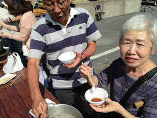 屋外のテーブルで男性が鍋から料理を取り分け、手前の女性が小皿に入ったタレと箸を手に笑顔を見せている様子の写真