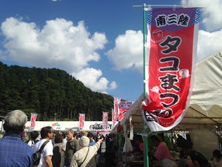 青空の下、「南三陸タコまつり」と書かれた赤いのぼりと白いテントの屋台が並び、多くの来場者で賑わうイベント会場の様子を撮影した写真