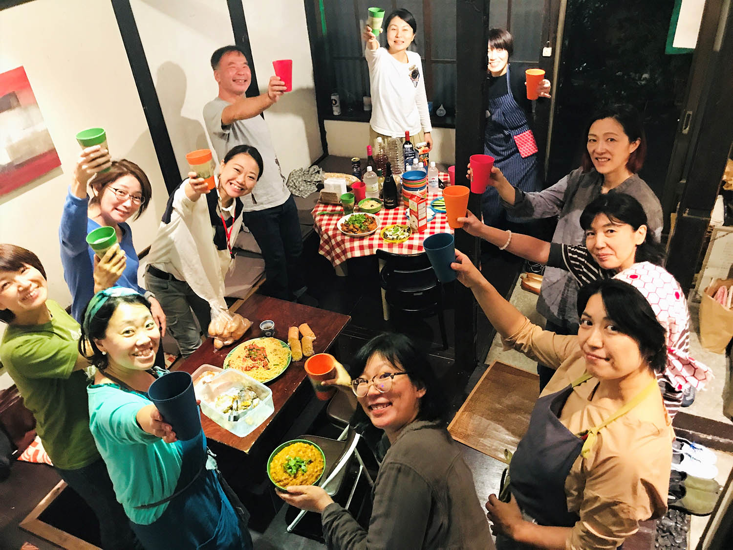 テーブルに料理や、おつまみが並べられ、パーティーに参加した方々が片手にコップを持って乾杯する楽しいそうな様子の写真