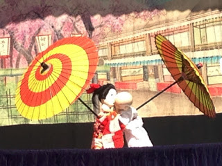 舞台上で赤と黄の和傘を持った着物姿の人形二体が抱き合い、背景に桜や町並みが描かれている人形劇の様子を撮影した写真