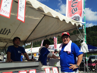 青空の下、白いテントの屋台で青いシャツを着た男性が笑顔で立ち、2人の男性が作業をしている写真