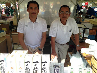 屋外テントの下で白いポロシャツを着た二人の男性が、日本酒「一ノ蔵」の箱や瓶が並ぶ販売ブースに立っている写真