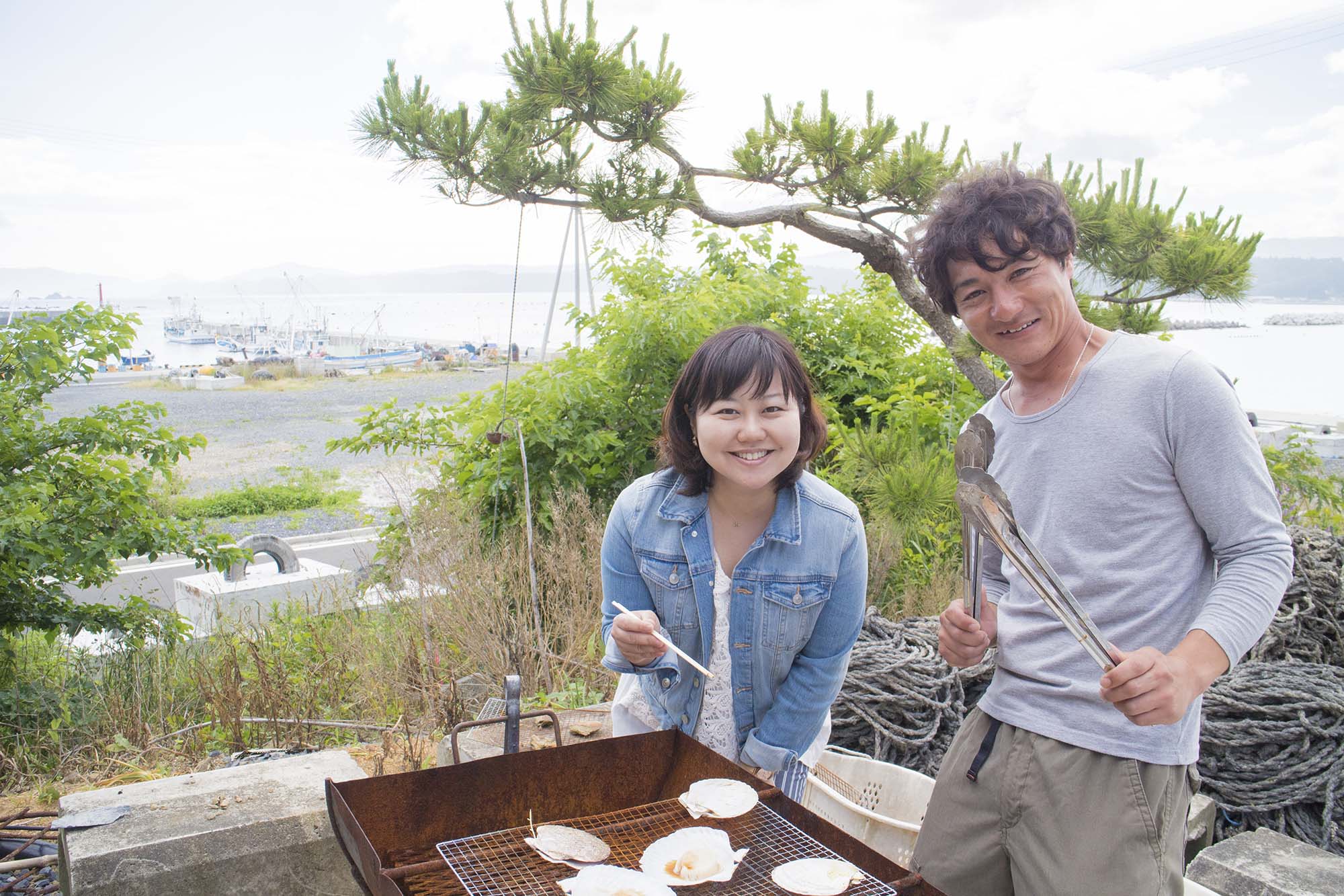 海を背景に、屋外に置かれたバーベキュー用の網の上に殻付きのホタテが並び、横に立った女性リポーターが箸、隣に立った男性がトングを持って写っている写真