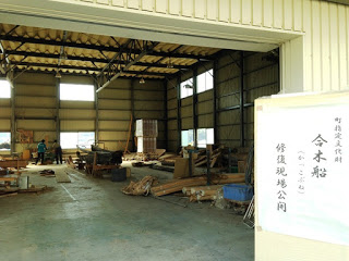 「合木船修復現場公開」と書かれた立て札のある建物の入り口から、中で木材や船が並ぶ修復作業場を見渡している写真
