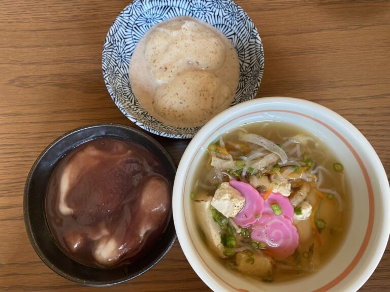 お椀に盛られた具だくさんの郷土料理で、ピンク色のナルトと刻みねぎがのった澄んだ汁のうどんと、あんこがたっぷりかかった柔らかい餅2個、そしてきな粉と黒蜜のかかった餅2個が並べられている、南三陸町の郷土料理の写真