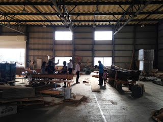 屋内作業場の奥で複数の人が古い木船の周りに集まり、作業や見学をしている様子を広く捉えた写真