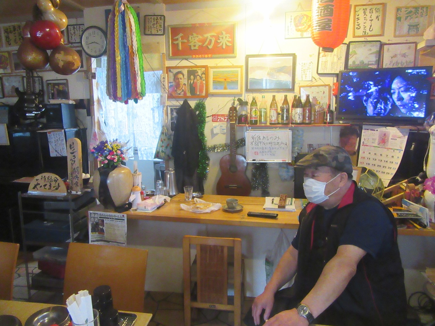 背景の壁にはお酒の瓶やテレビ、様々な写真や絵、飾りが所狭しと並んでいる温かみのある店内に、マスク姿でカウンター横に座っている店主の渡辺さんの写真