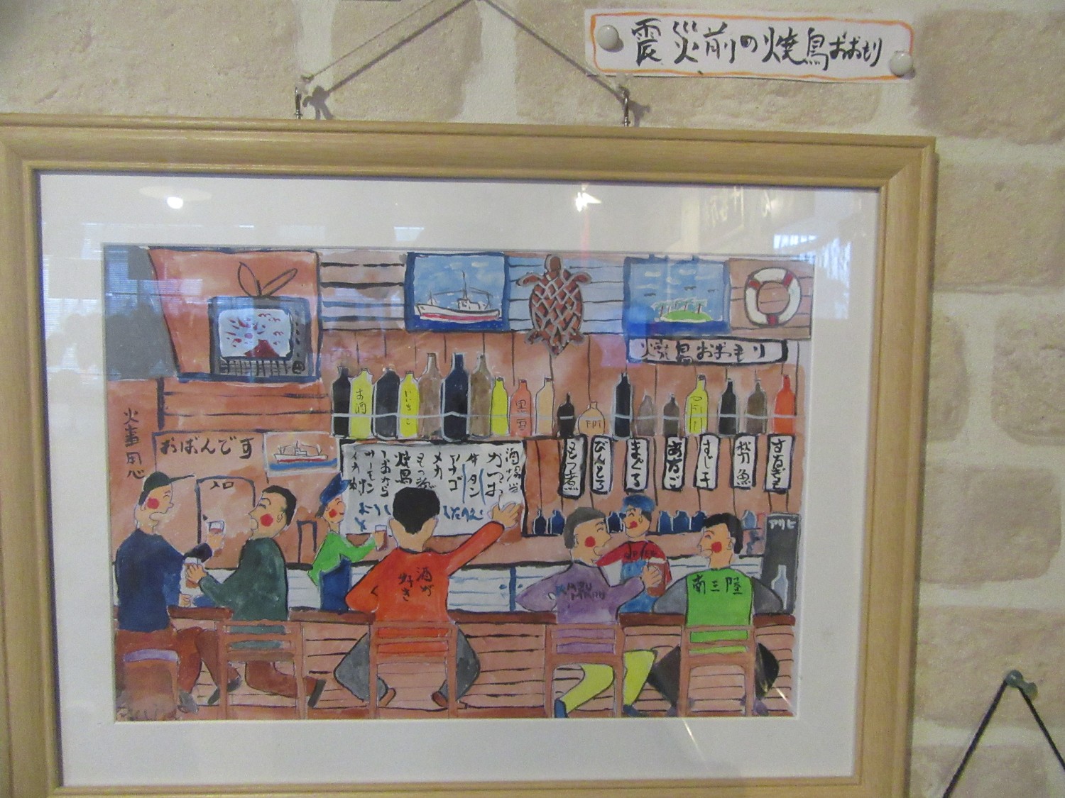 背景には酒瓶とメニューが並ぶ棚があり、カウンターのある居酒屋のような店内で数人の客が酒を飲みながら談笑している様子が描かれている色彩豊かな水彩画が額に入れられ、「震災前の焼鳥おおもり」と手書きされた紙と共に壁に掲げられている写真