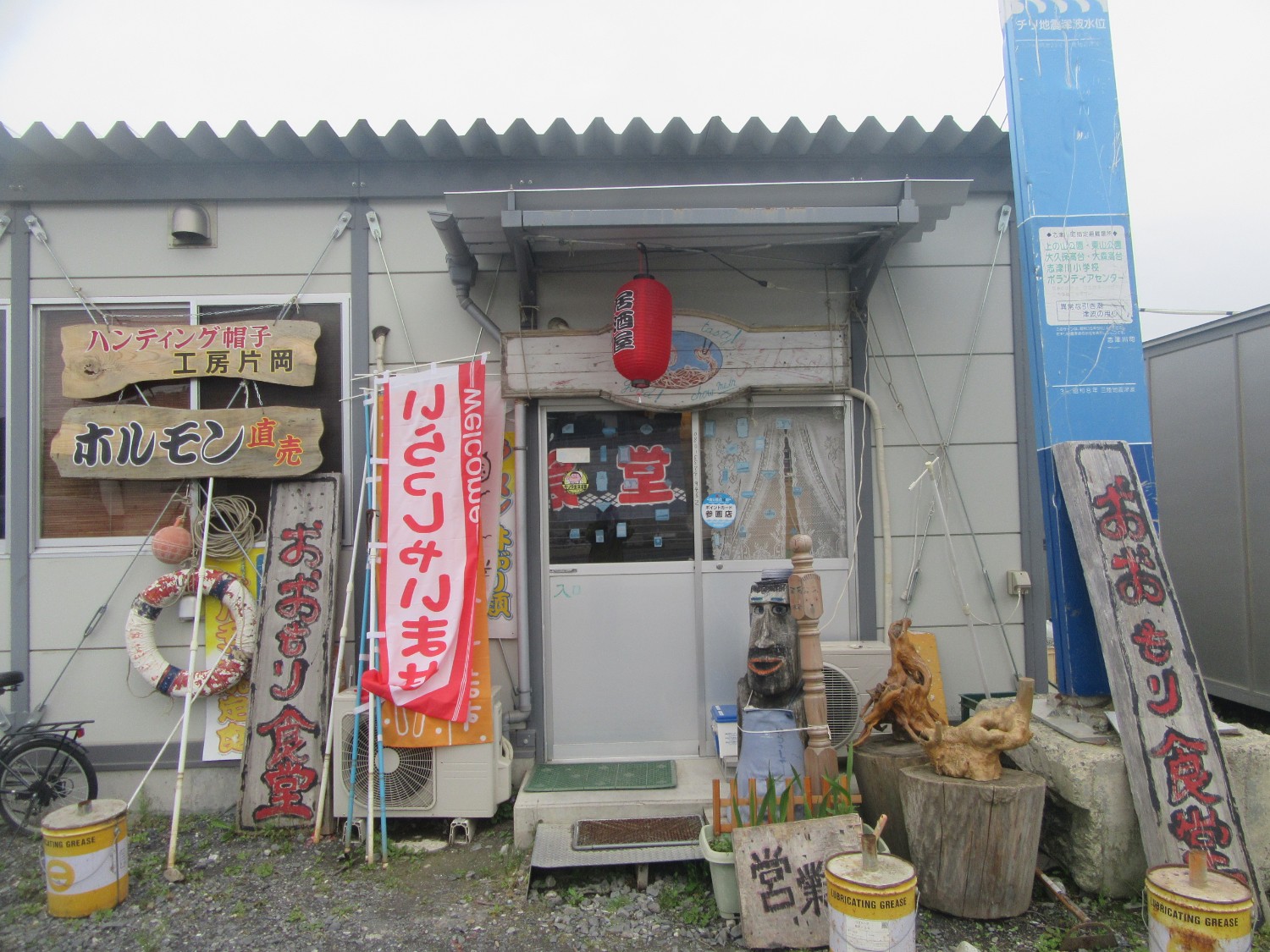 周囲に木彫りのオブジェやのぼり旗、手書きの営業案内などが立てかけられているプレハブ風の建物の正面に「おおもり食堂」と書かれた木の看板や赤ちょうちんがある、小さな食堂の入口の写真