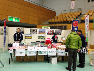 屋内ブースにて地元特産品を販売するテーブルの奥に立つ出店者と、それを見ている来場者が写っている写真