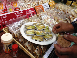 試食用の皿に並んだ焼き野菜のような料理が中央に置かれ、周囲には煎餅などのパッケージ商品が並んでいる写真