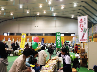 体育館のような屋内会場で、さまざまな野菜や加工品のブースが並び、多くの来場者が買い物や見学をしている写真