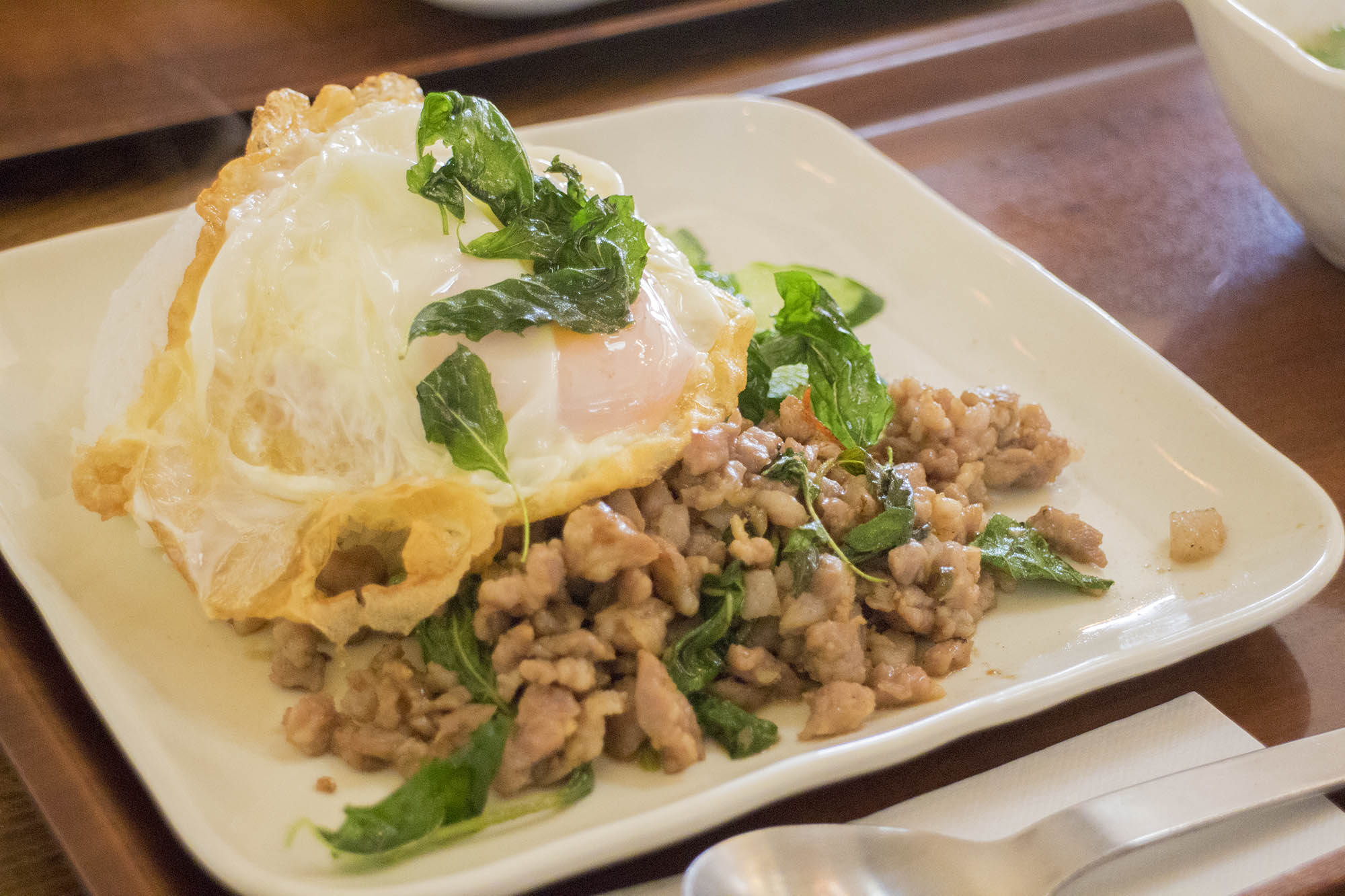 白い四角い皿に、バジルと炒めたひき肉の上にカリッと揚げた半熟目玉焼きがのせられた、タイ風の料理が盛りつけられている写真