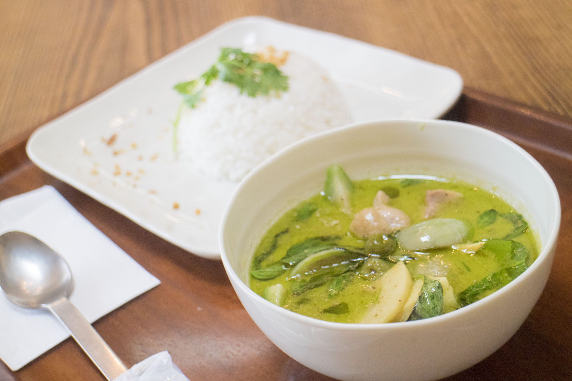 白いボウルに入った鶏肉やナス、たけのこなどが入ったタイ風グリーンカレーと、香草が添えられた白いご飯が四角い皿に盛られて一緒にトレーにのせられている写真