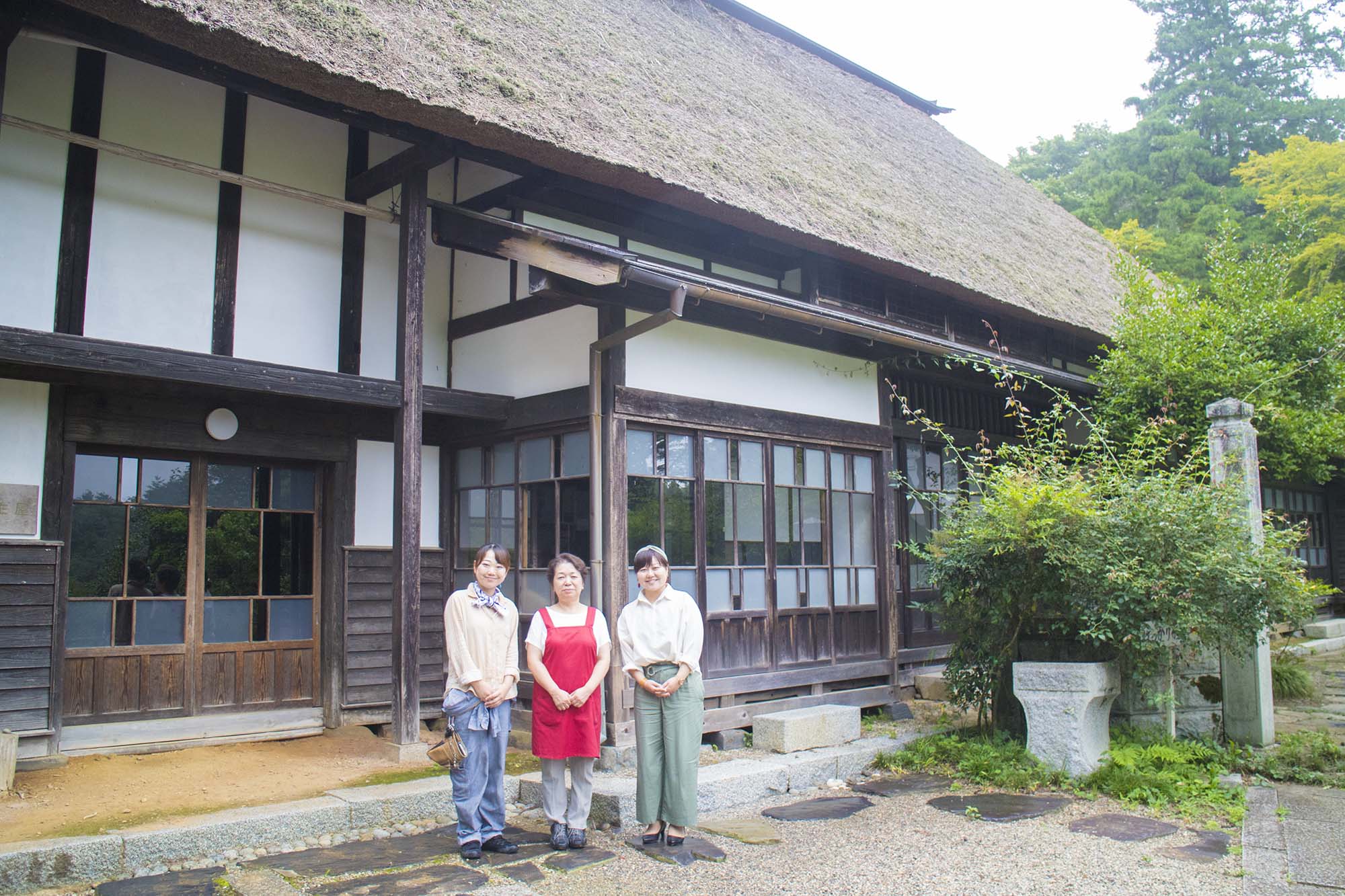 古い木造で茅葺屋根の日本家屋の前で、赤いエプロン姿の女性を中央に、ジーンズとベージュの長袖を着た女性と、白いシャツと緑のパンツ姿のリポーターの女性がそれぞれ左右に立ち、3人でカメラに向かって微笑んでいる写真