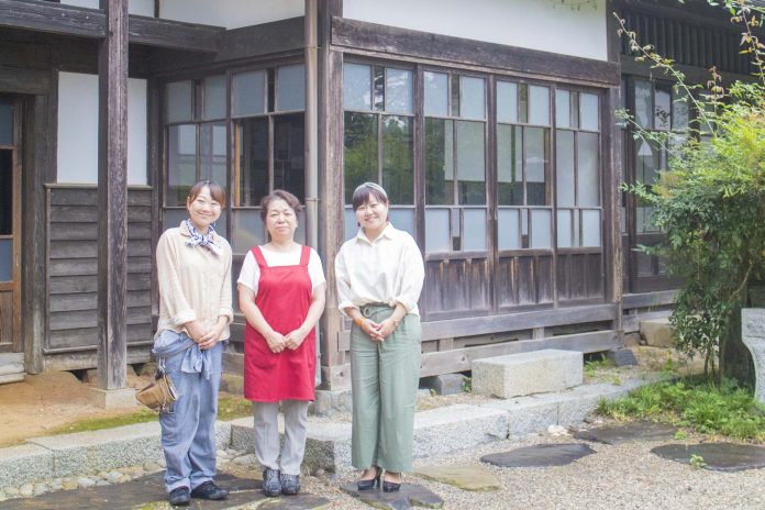 古い木造の日本家屋の前で、赤いエプロン姿の女性を中央に、ジーンズとベージュの長袖を着た女性と、白いシャツと緑のパンツ姿のリポーターの女性がそれぞれ左右に立ち、3人でカメラに向かって微笑んでいる写真