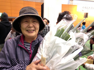 帽子をかぶった高齢の女性が、購入したたくさんの長ネギを笑顔で両手に抱えている写真