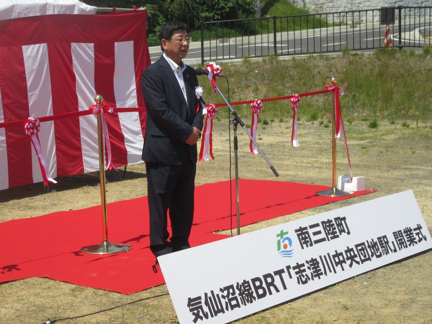 背景には紅白幕が貼られ「南三陸町 気仙沼線BRT『志津川中央団地駅』開業式」と書かれた看板の後ろに敷かれたレッドカーペットの上でマイクスタンドに向かって立った佐藤町長が挨拶をしている様子の写真