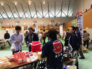 タコの絵が描かれた法被を着た男性たちが、屋内のブースでお菓子などの商品を販売し、来場者と会話している写真