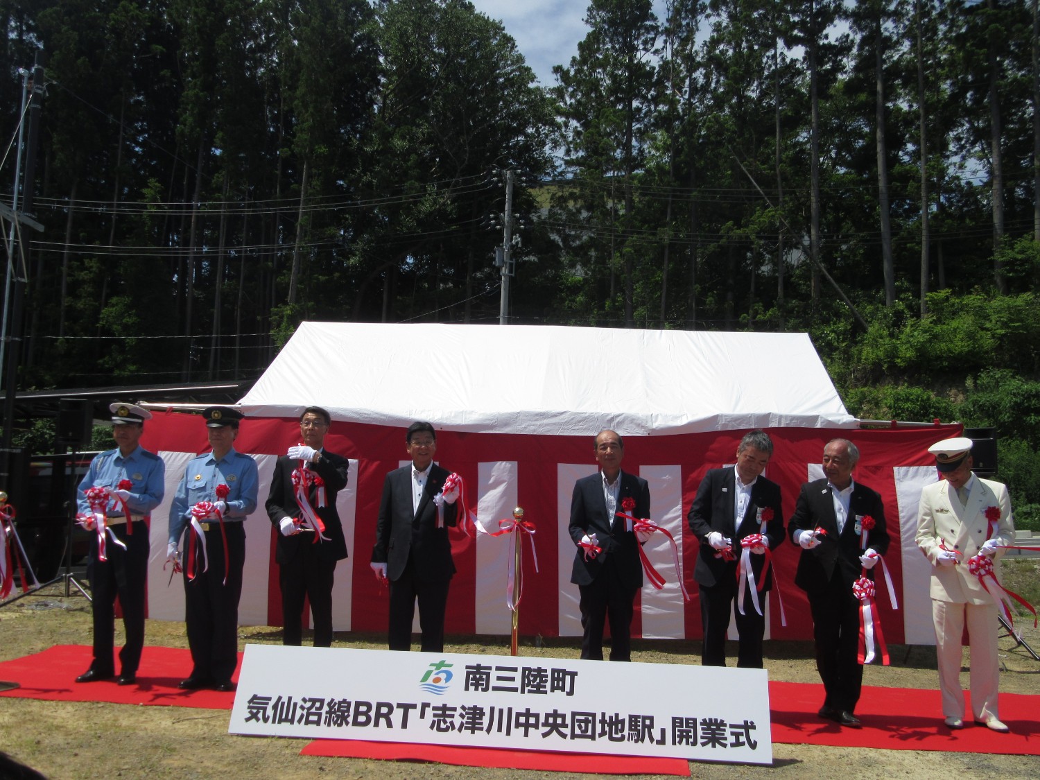 背景には紅白幕が貼られ「南三陸町 気仙沼線BRT『志津川中央団地駅』開業式」と書かれた看板の後ろに、警察官やスーツ姿の関係者たちが並んで立ち、テープカットが行われている写真