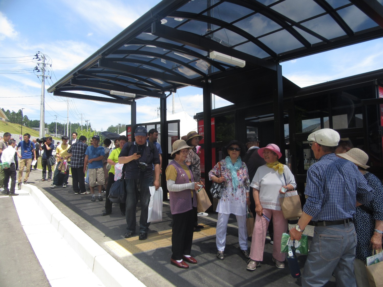 取材カメラが撮影している新しいバス停のアーチ型の黒い屋根の下に、多くの人々が楽し気に立っている写真
