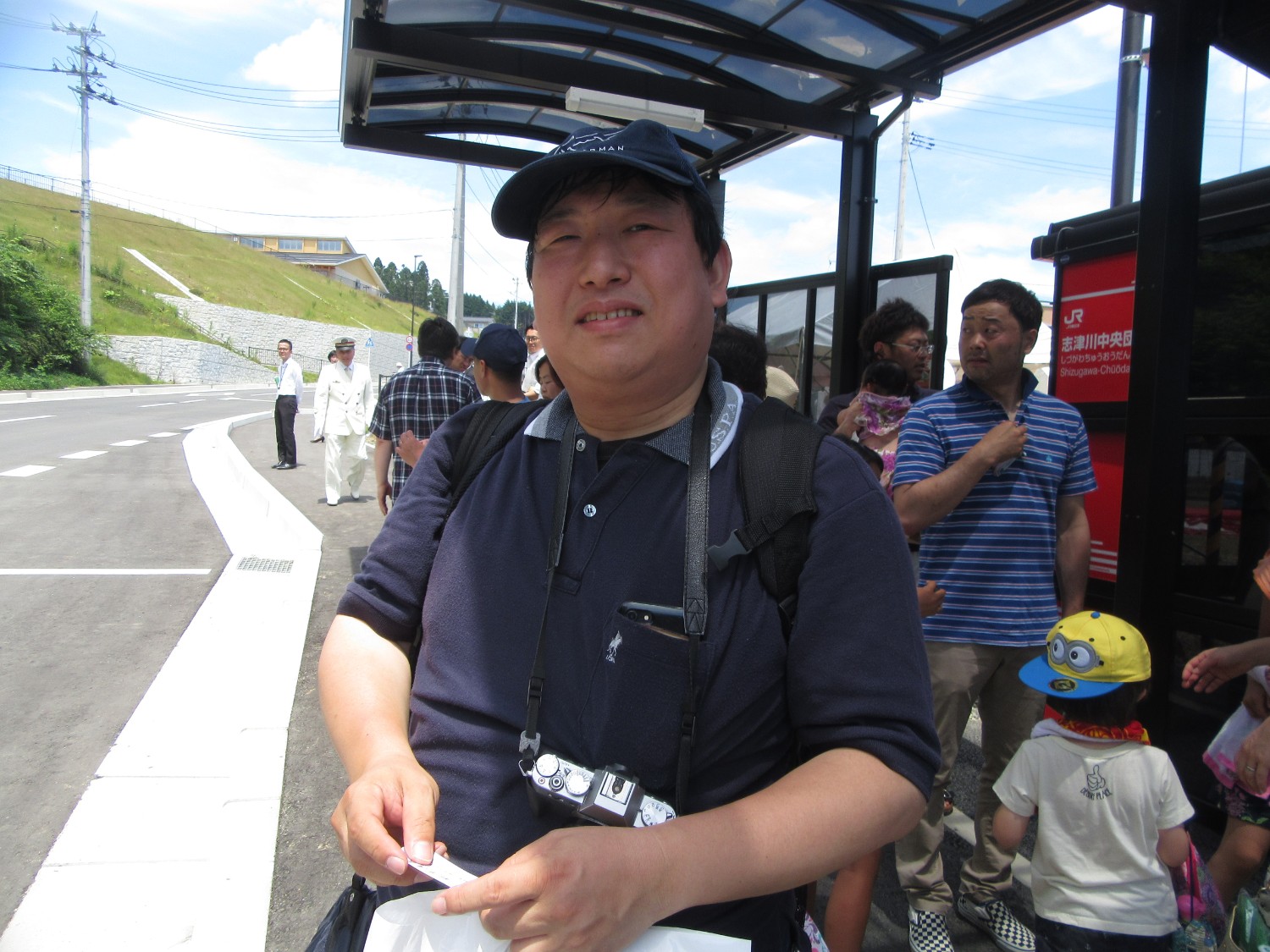 バス停に並んでいる、カメラを首から下げ帽子を被った男性が手にチケットのようなものを持ってカメラに笑顔を見せている写真
