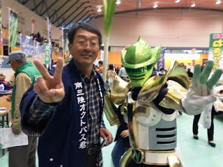 「南三陸オクトパス君」と書かれた法被を着た男性と、緑色の戦隊ヒーロー風の着ぐるみキャラクターが並んでピースサインをしている写真