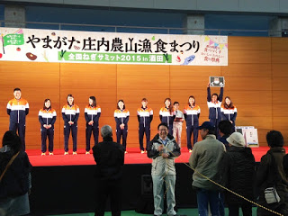 ステージ上でジャージ姿の若者たちが整列し、後方に「やまがた庄内農山漁食まつり」と書かれた横断幕が掲げられている写真