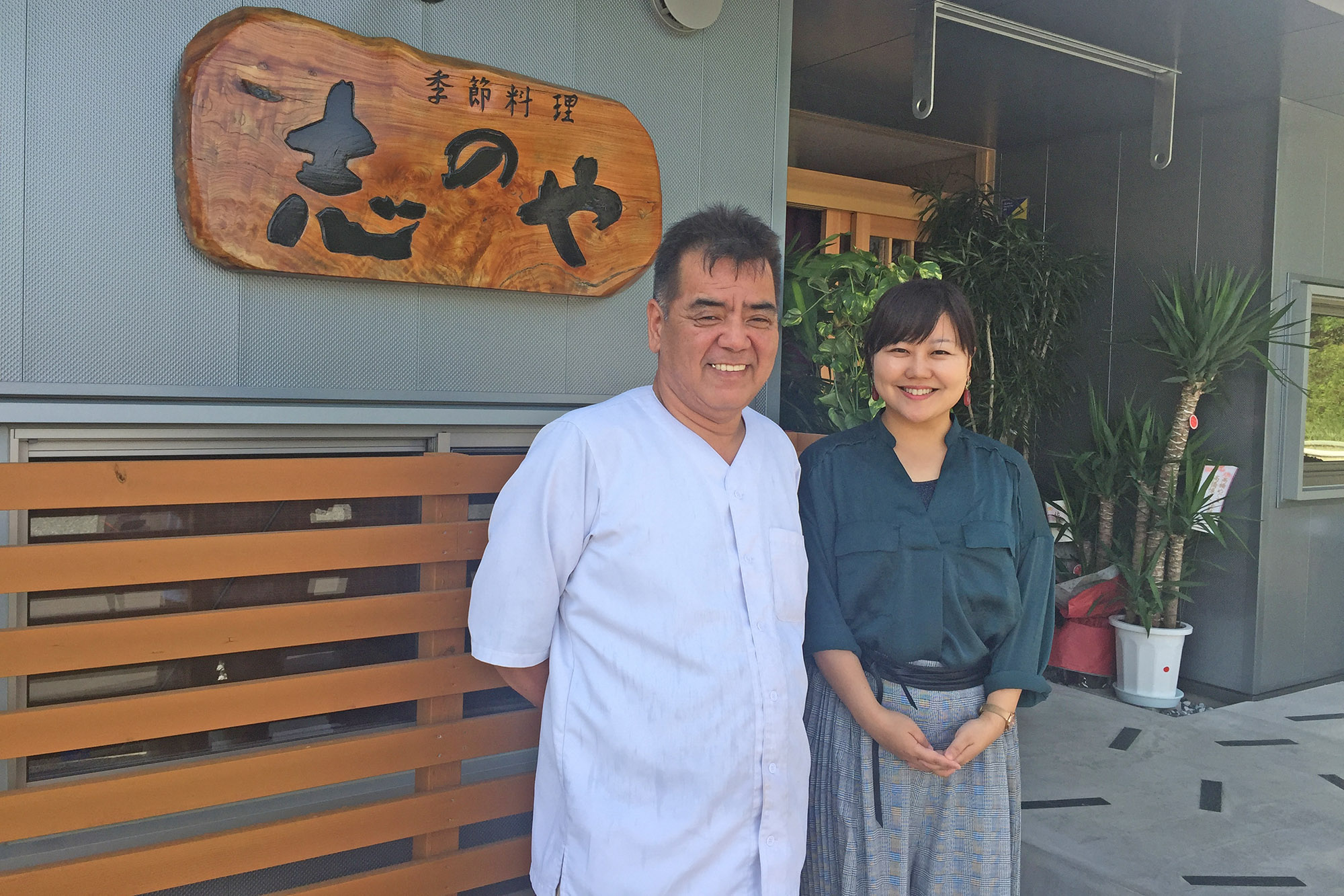 木製の看板に「季節料理 志のや」と書かれた店舗の前で、白衣を着た料理人の高橋さんと女性リポーターが並んで笑顔で立っている写真