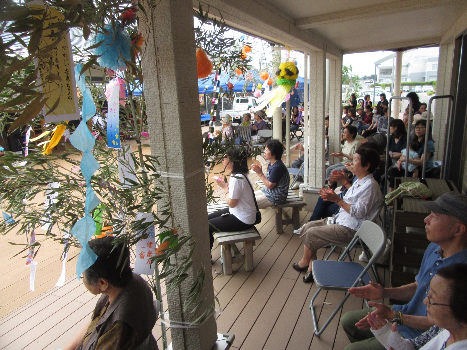 バルコニーの柱に取り付けた笹の葉に短冊や飾りが吊るされ、その向こうに多くの観客がベンチや椅子に座って演目を見て拍手している様子の写真