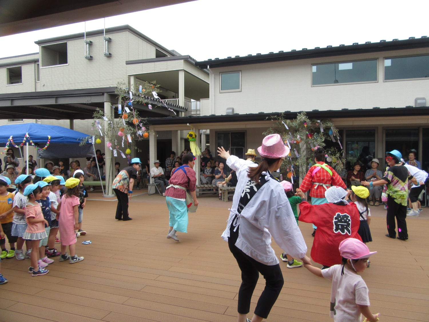 園児たちと割烹着や着物を着たスタッフ、踊り手たちが中庭の広場で一緒に踊りを楽しんでいる様子の写真