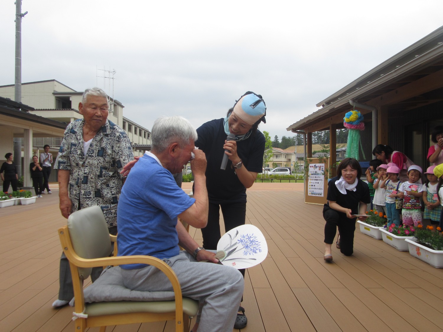 うちわを持ち椅子に座る高齢の男性が目元をぬぐっており、ちょんまげのかつらをかぶりマイクを持っている女性が背中をやさしくなで、周囲にいる人たちが温かく見守っている写真